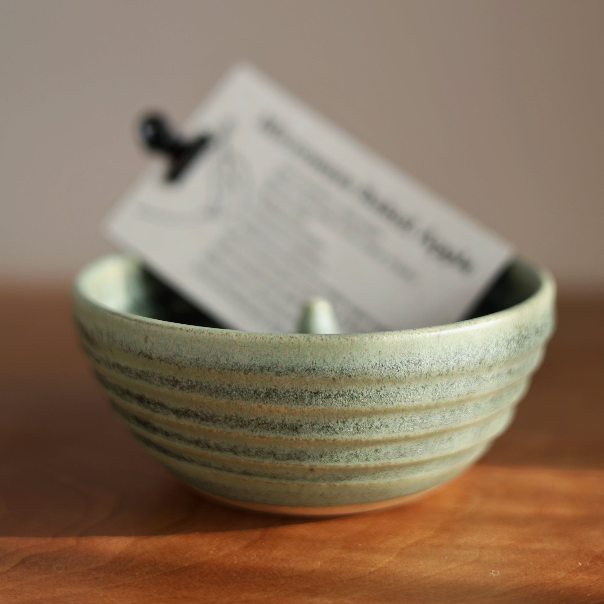A small ribbed ceramic apple baker bowl in a pale moss green glaze sits on a wooden surface with a white paper tag tucked inside, photographed against a soft neutral background.