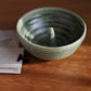 A handmade ceramic apple baker with a textured, speckled sage green glaze, shown on a wooden table next to printed cards from 'From the Gravesco Kitchen'.