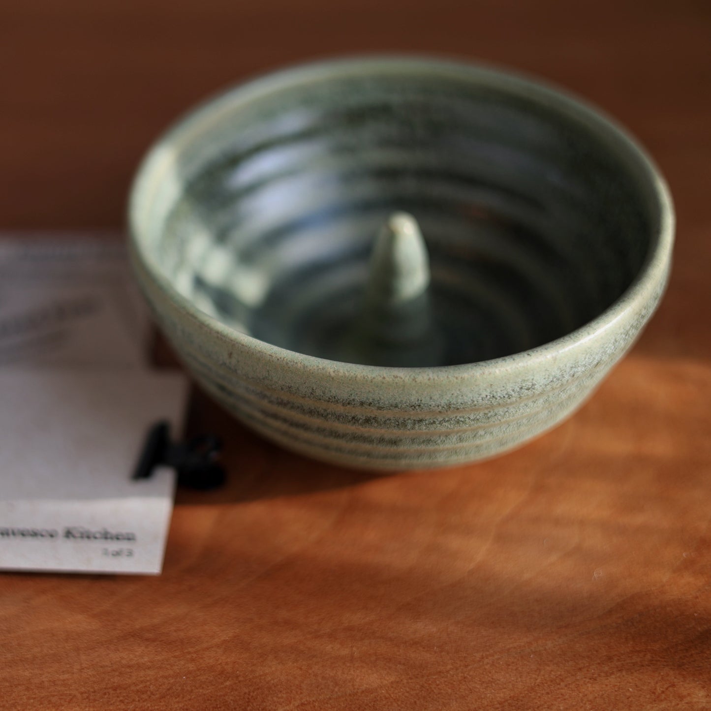 A handmade ceramic apple baker with a textured, speckled sage green glaze, shown on a wooden table next to printed cards from 'From the Gravesco Kitchen'.