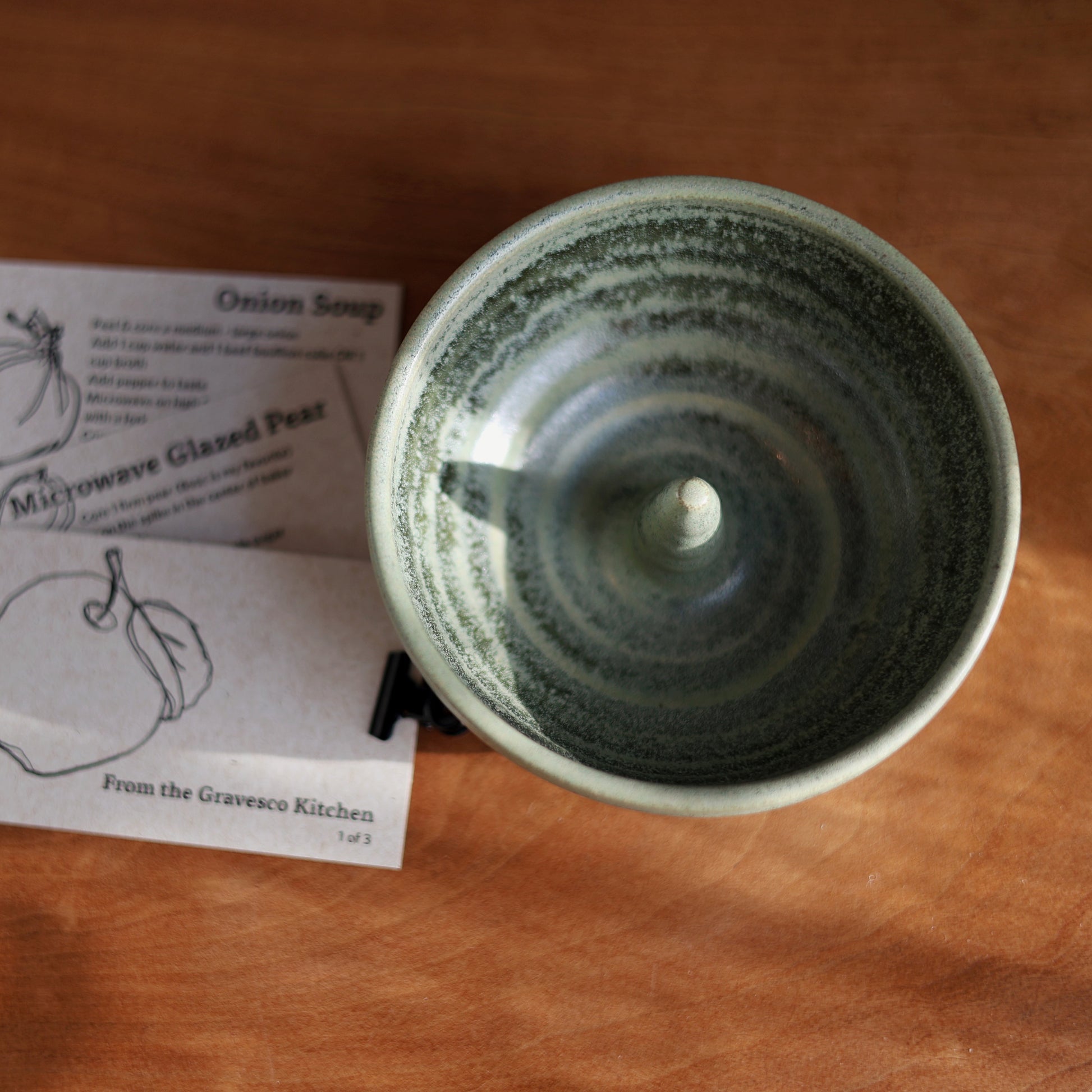 A handmade ceramic bowl with a speckled green glaze sits on a wooden table next to two printed recipe cards titled 'Onion Soup' and 'Microwave Glazed Pear' from 'From the Gravesco Kitchen'.