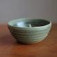 A small handmade ceramic apple baker with a speckled sage green glaze and horizontal ridges, sitting on a wooden tabletop against a neutral gray background.