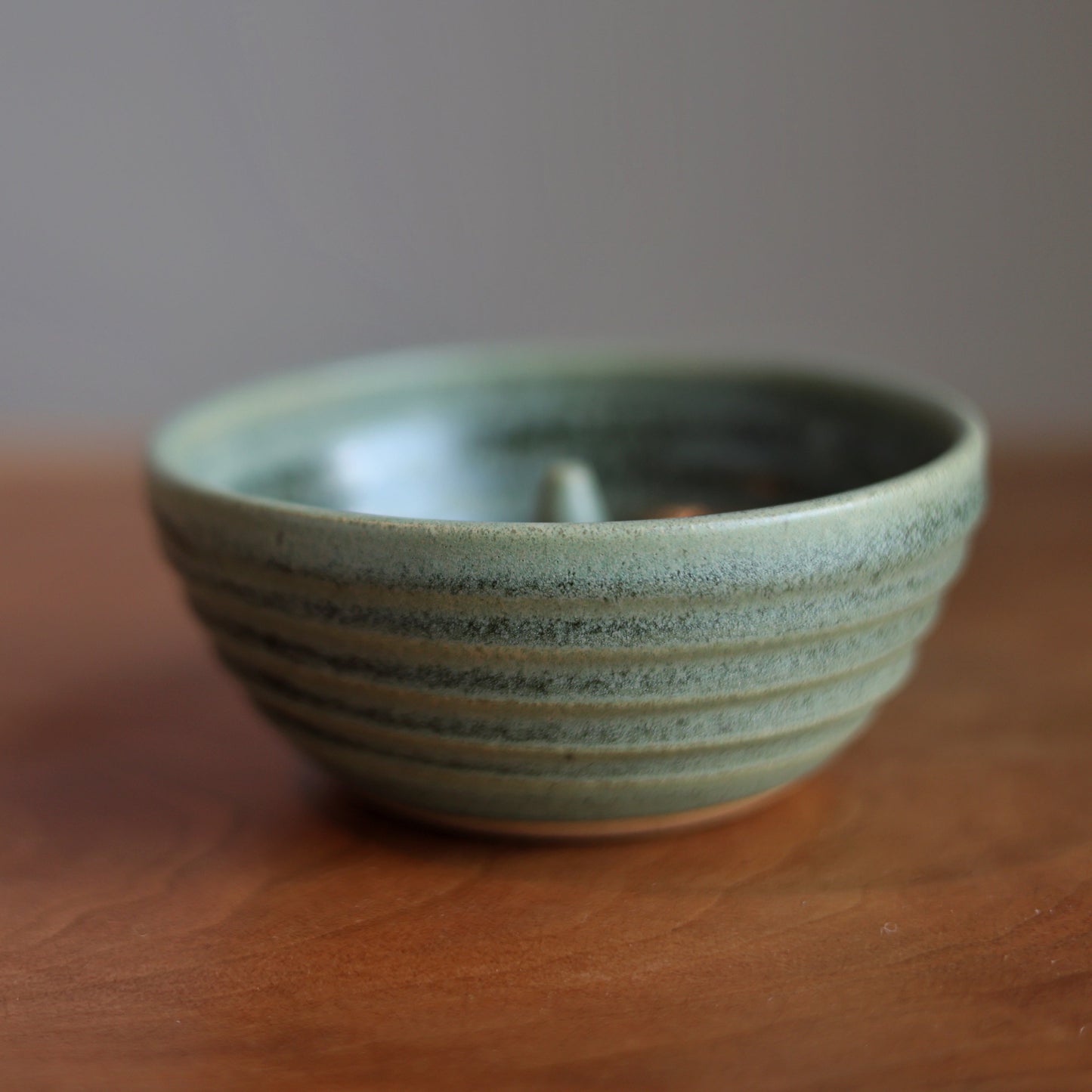A small handmade ceramic apple baker with a speckled sage green glaze and horizontal ridges, sitting on a wooden tabletop against a neutral gray background.