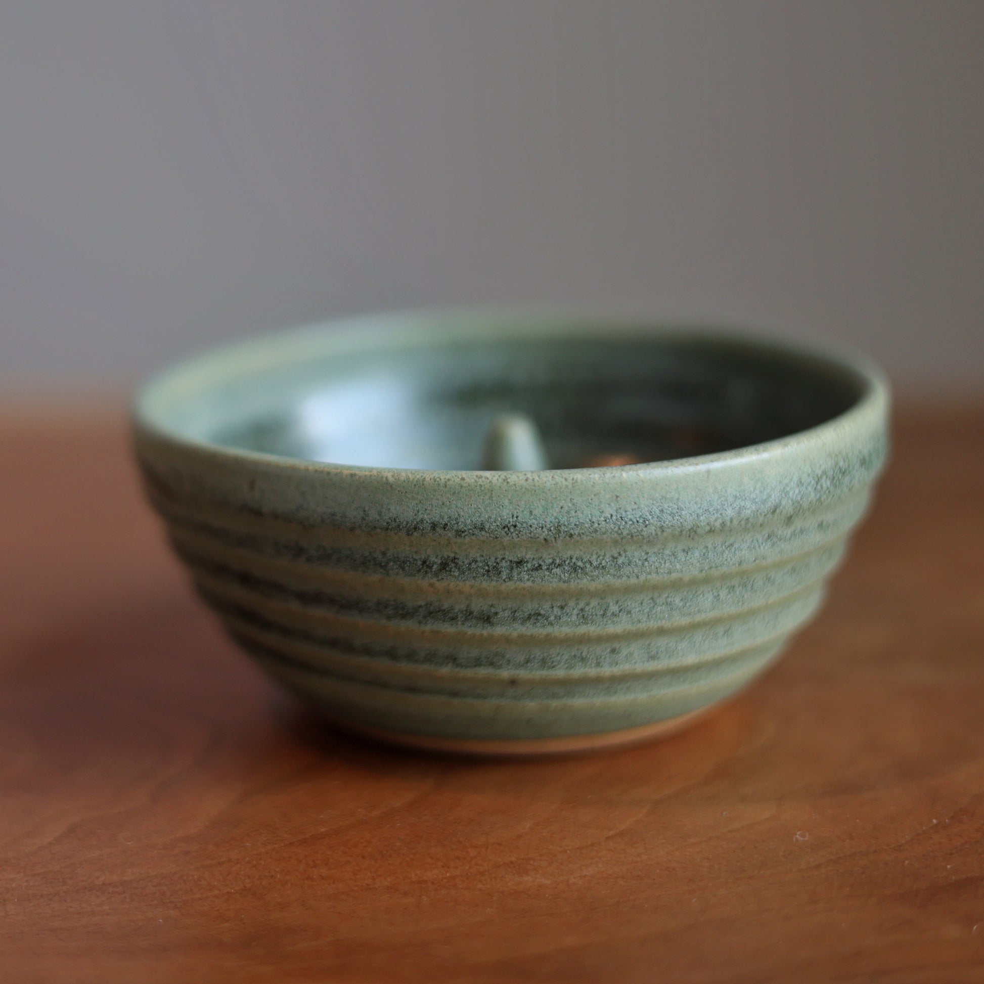 A small handmade ceramic apple baker with a speckled sage green glaze and horizontal ridges, sitting on a wooden tabletop against a neutral gray background.