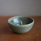 A handmade ceramic bowl with a textured, ribbed exterior and a glossy, light green glaze, resting on a wooden surface against a neutral gray background.