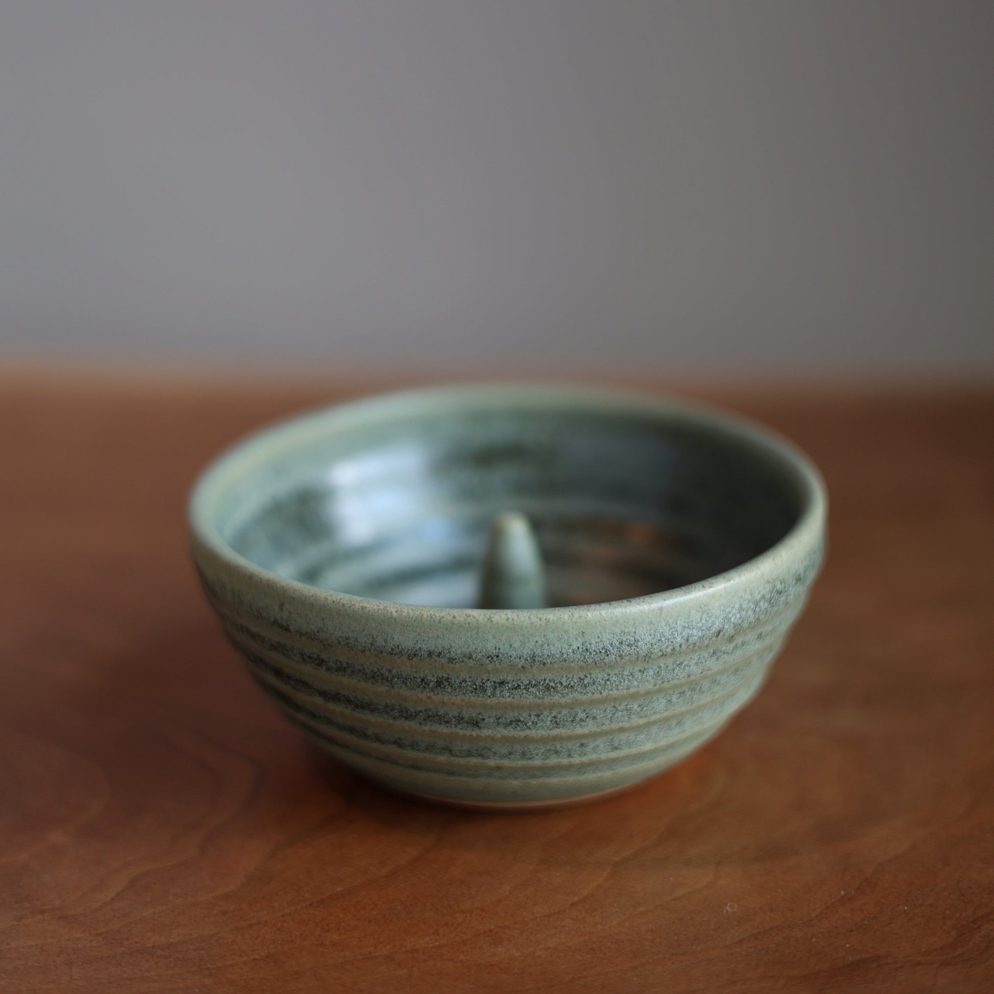 A handmade ceramic bowl with a textured, ribbed exterior and a glossy, light green glaze, resting on a wooden surface against a neutral gray background.