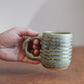 Hand holding a moss green ceramic mug with a striped pattern on a wooden surface.