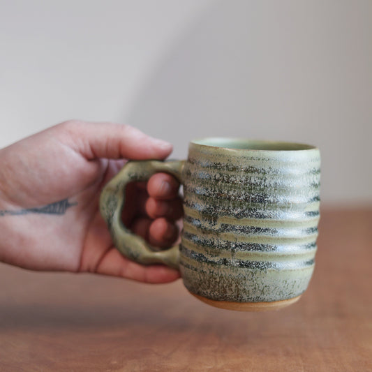 Hand holding a moss green ceramic mug with a striped pattern on a wooden surface.