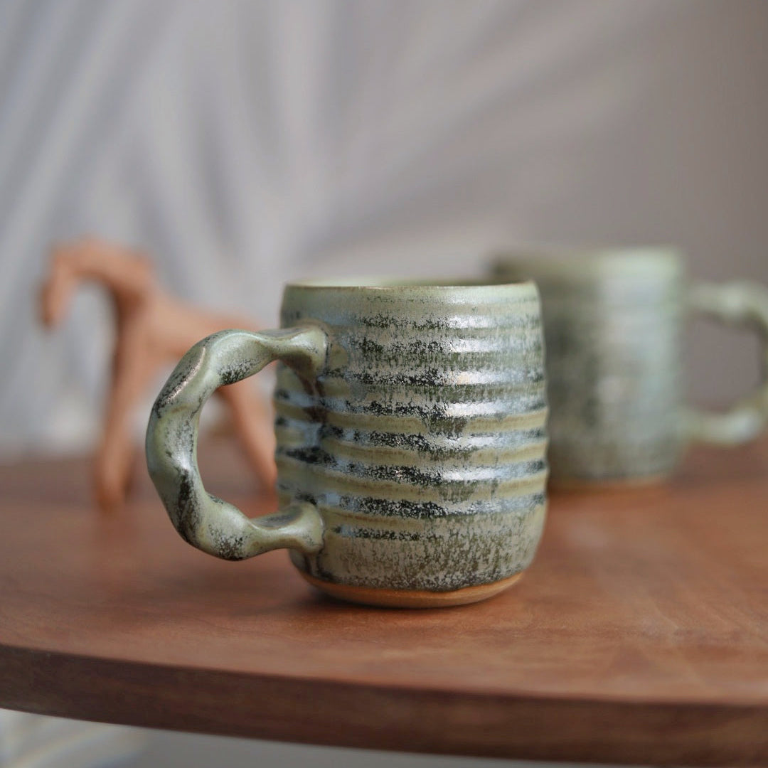 Two ceramic handmade moss green companion mugs on a wooden surface with a blurred background