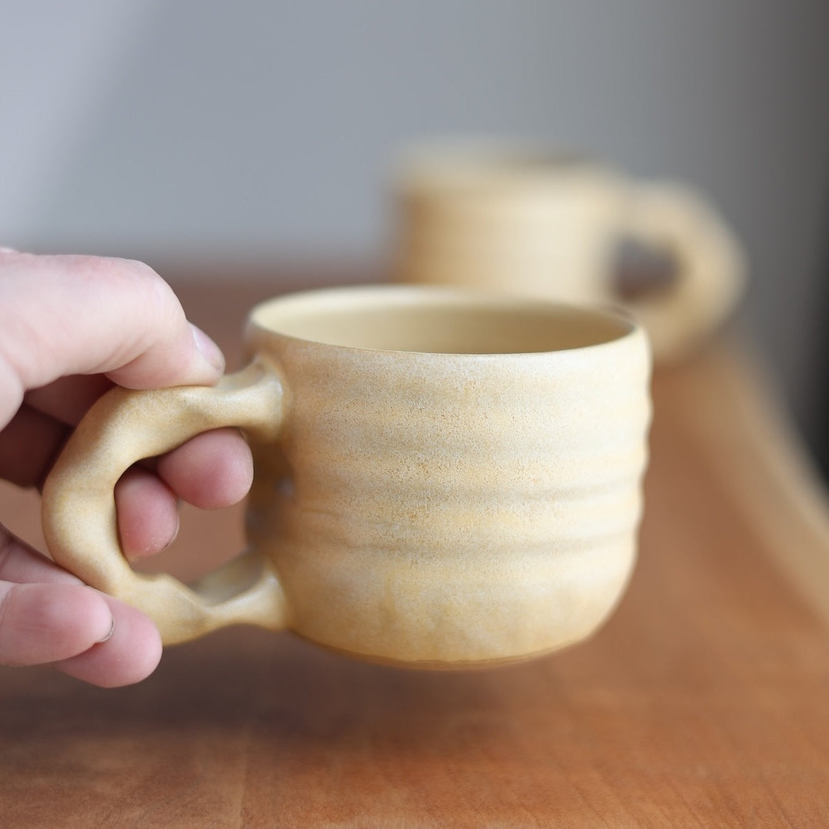Hand holding a ceramic mug on a wooden surface with another mug in the background.