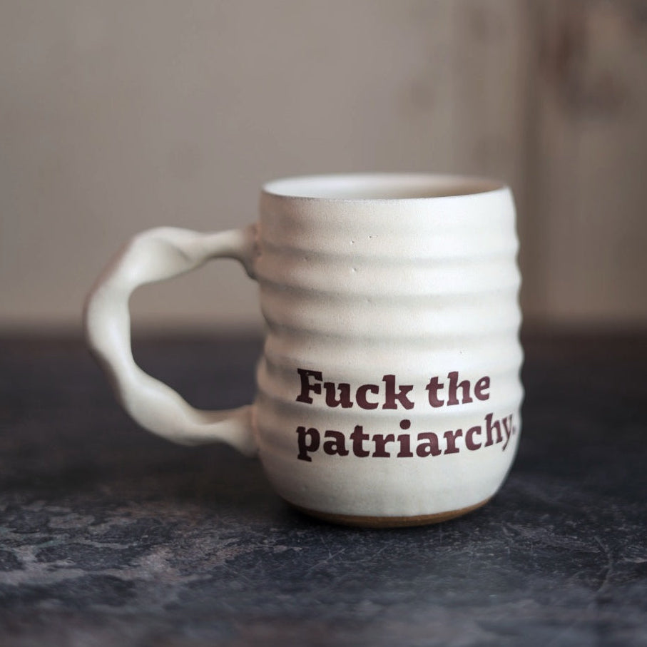 White ceramic mug with 'Fuck the patriarchy' text on a dark surface.