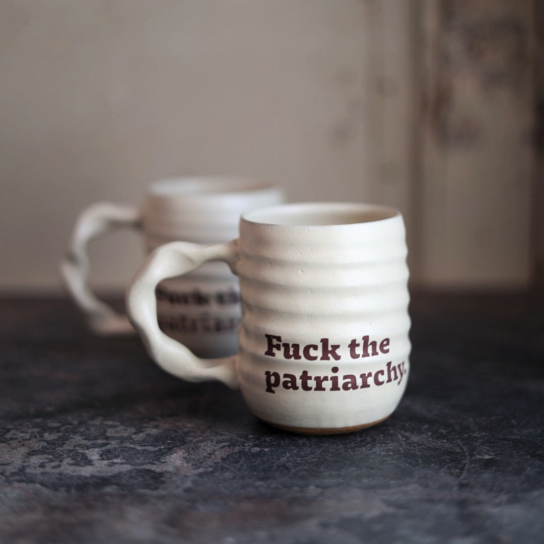 White ceramic mug with 'Fuck the patriarchy' text on a dark surface.