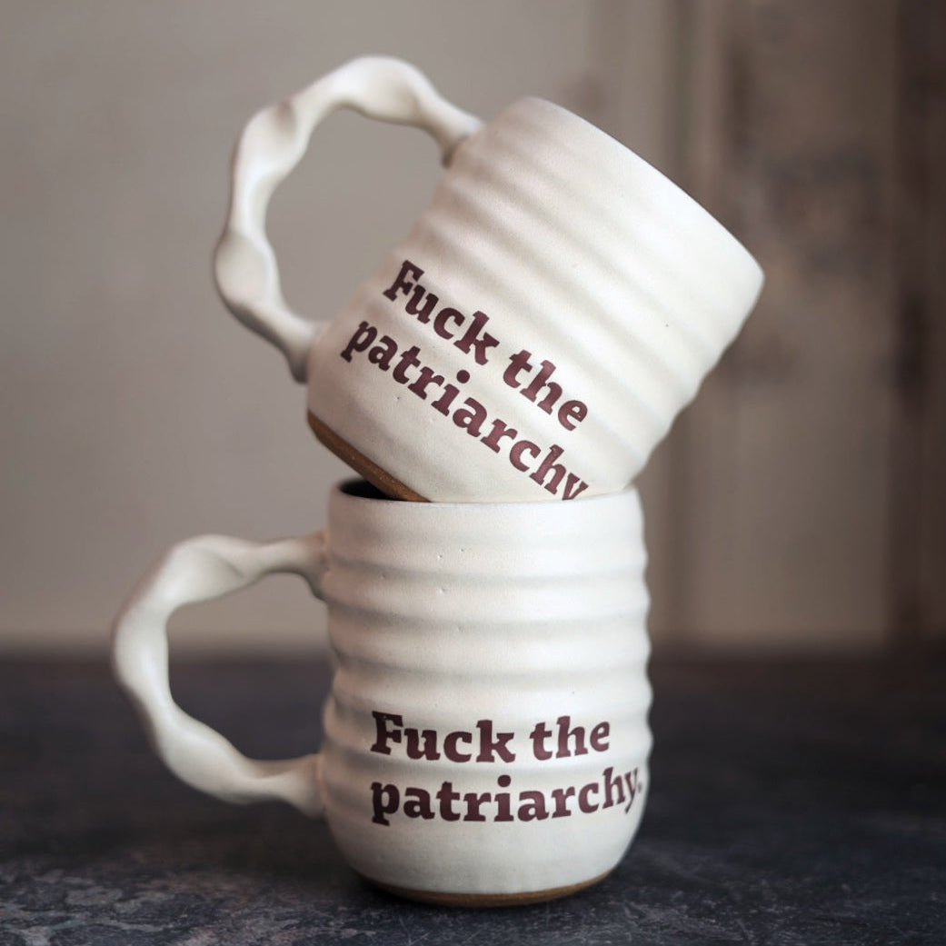Two white mugs with 'Fuck the patriarchy' text stacked on a dark surface.