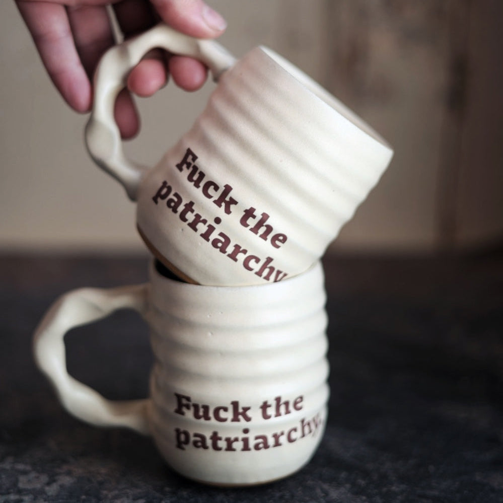 Two stacked ceramic mugs with 'Fuck the patriarchy' text on a dark surface.