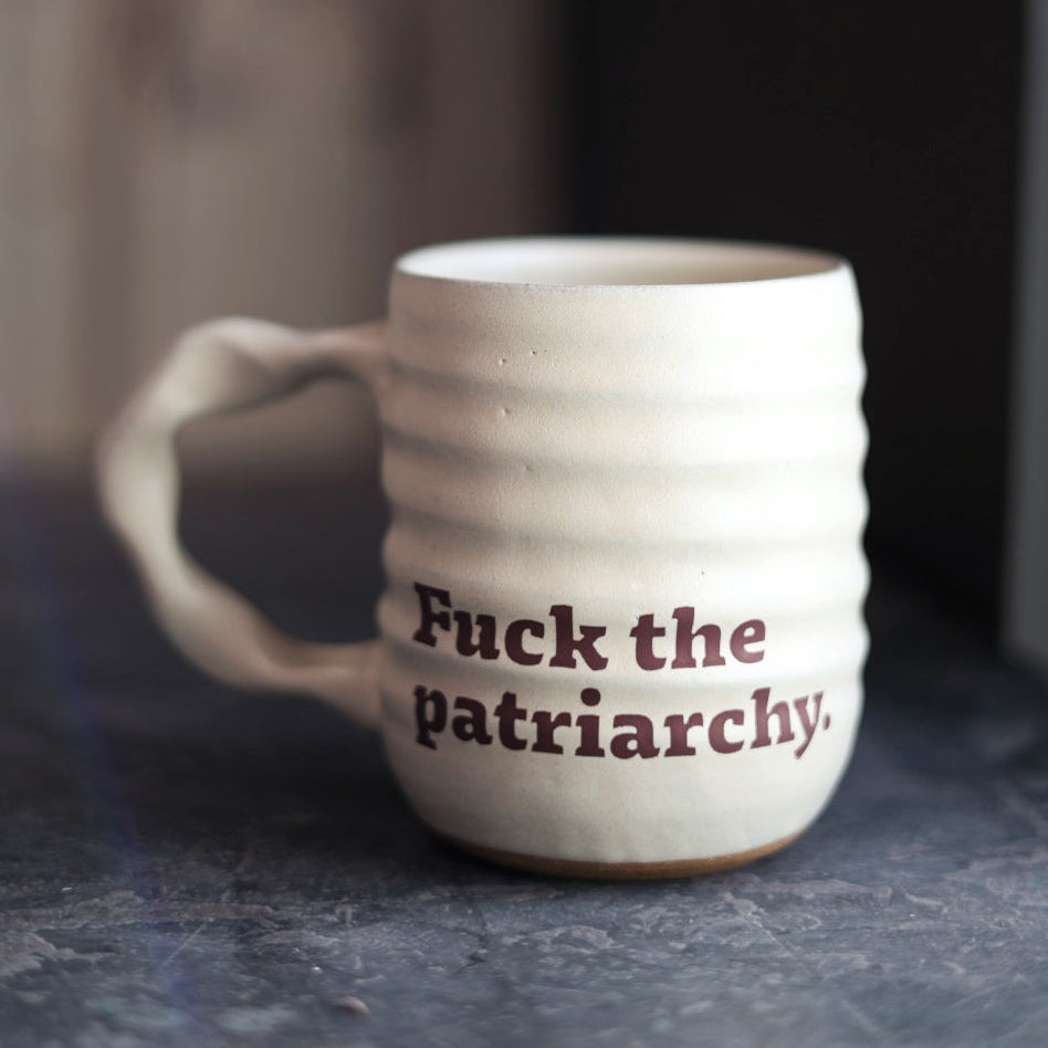 White ceramic mug with 'Fuck the patriarchy.' text on a dark surface.