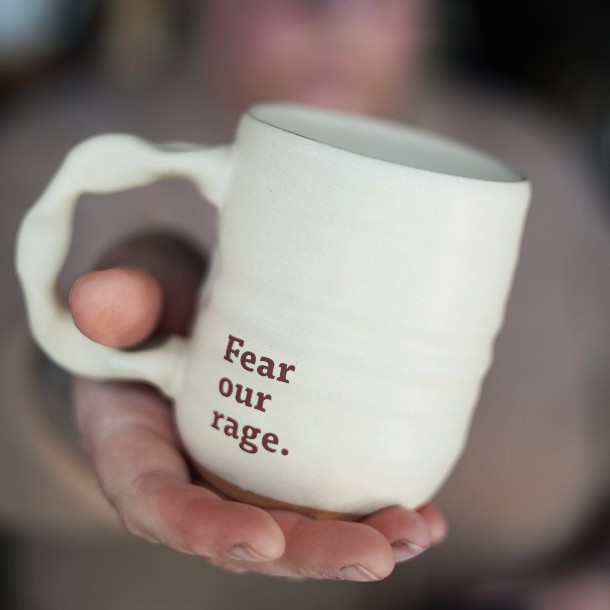 Person holding a mug with 'Fear our rage.' text, blurred background