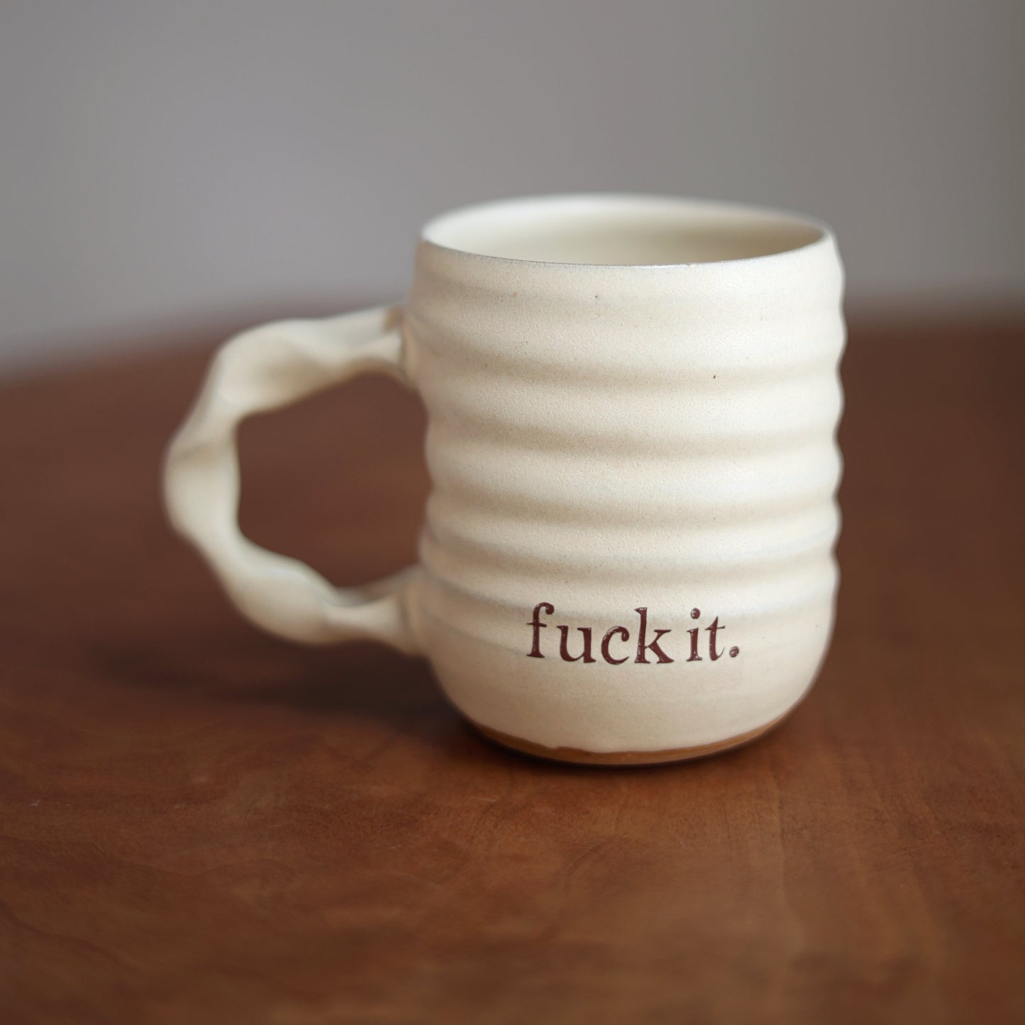 Cream ceramic mug with 'fuck it.' text on a wooden surface and gray background