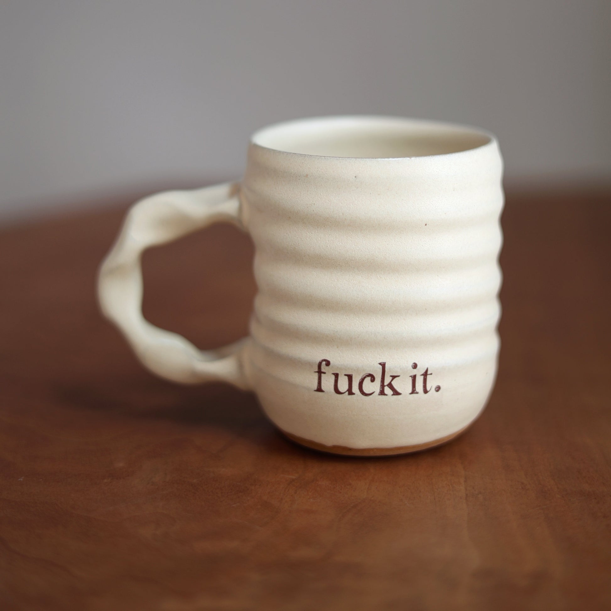 Cream ceramic mug with 'fuck it.' text on a wooden surface and gray background