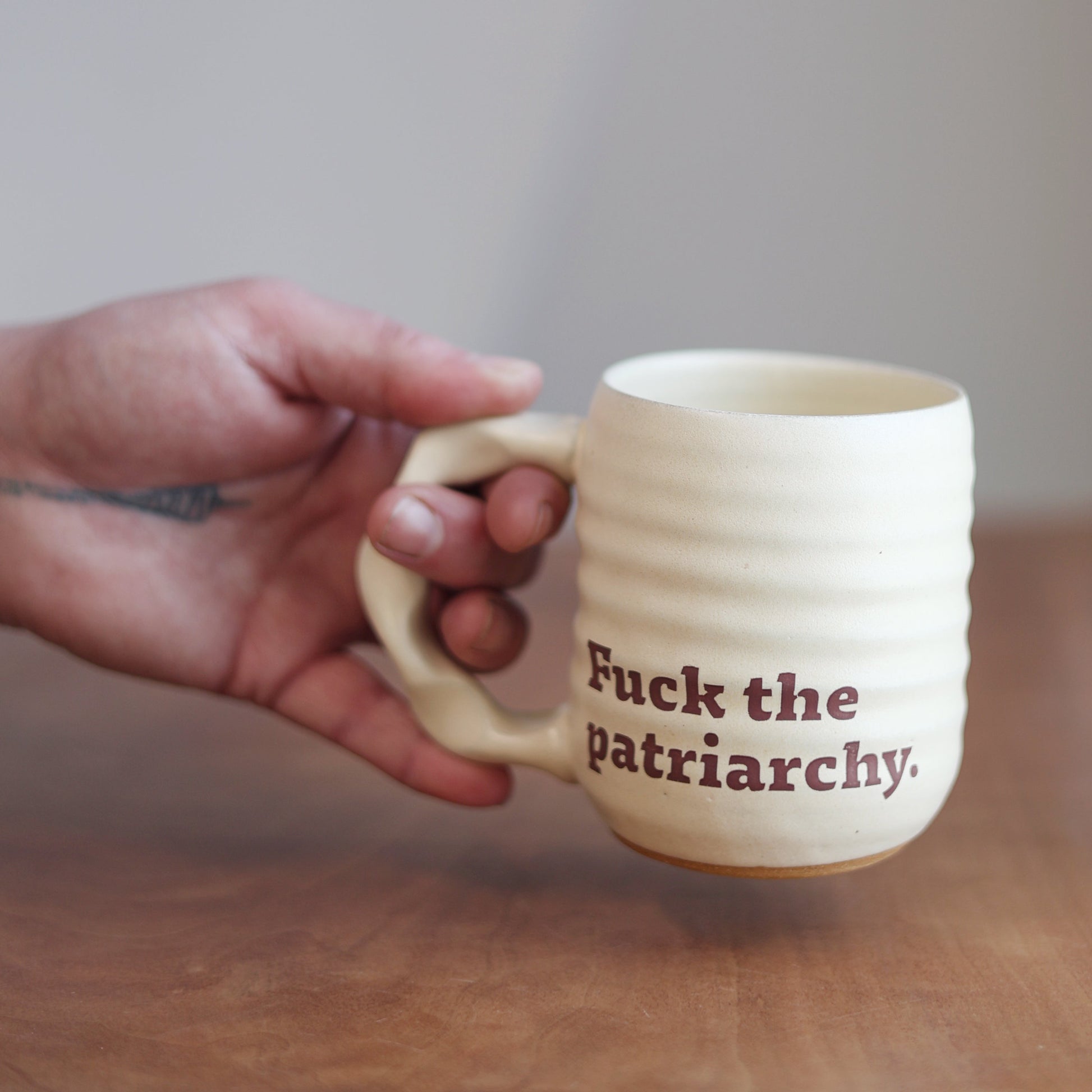 Hand holding a mug with 'Fuck the patriarchy' text on a wooden surface.