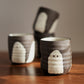 Three dark brown and white ceramic cups with a ghost design, arranged on a wooden table against a neutral gray background.