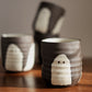 Three handmade dark brown and white ceramic cups with a ghost design, arranged on a wooden table against a neutral background.