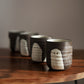 Four dark brown ceramic cups with white ghost-like faces, arranged in a diagonal row on a wooden table against a neutral gray background.
