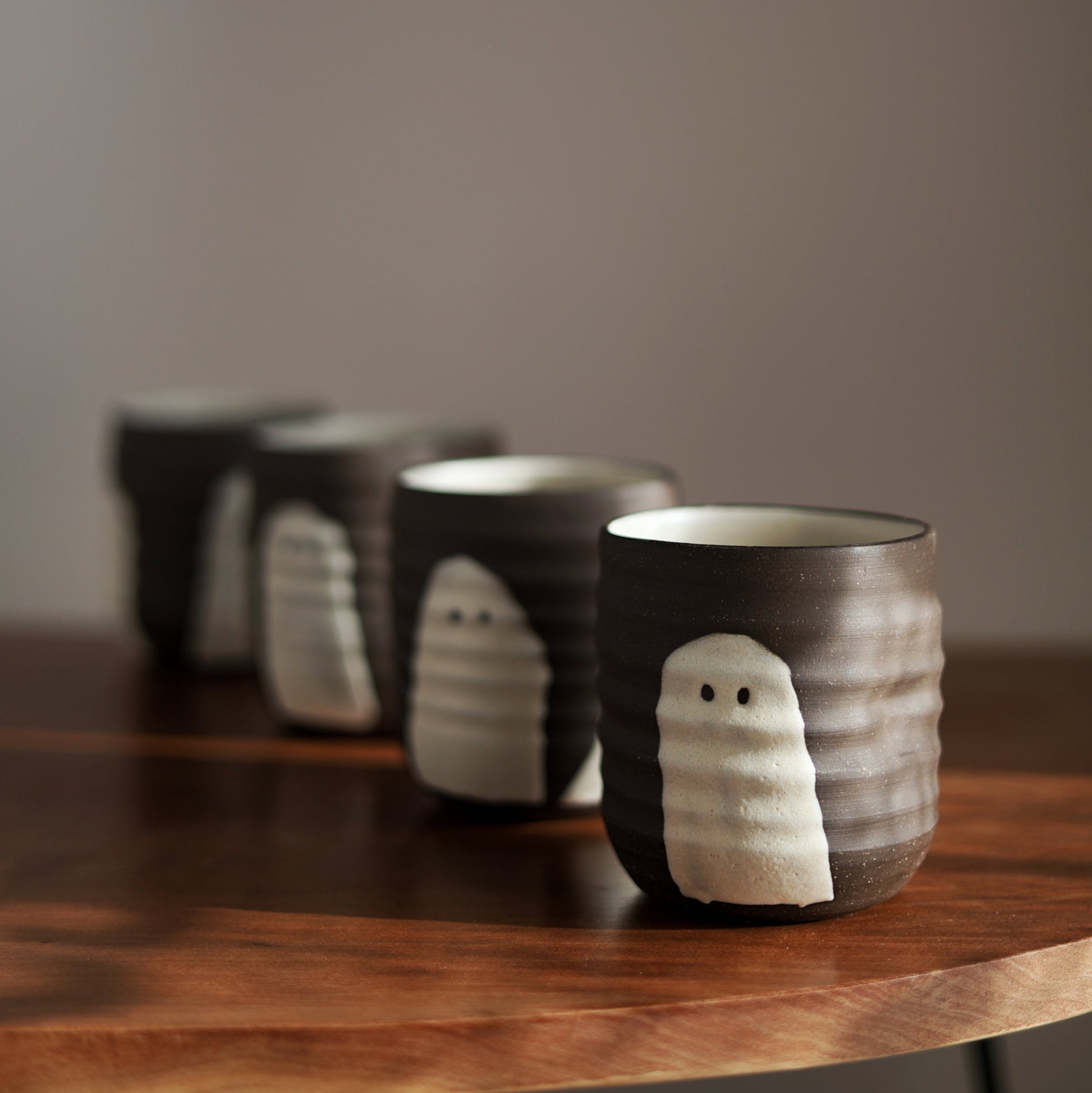 Four dark brown ceramic cups with white ghost-like faces, arranged in a diagonal row on a wooden table against a neutral gray background.