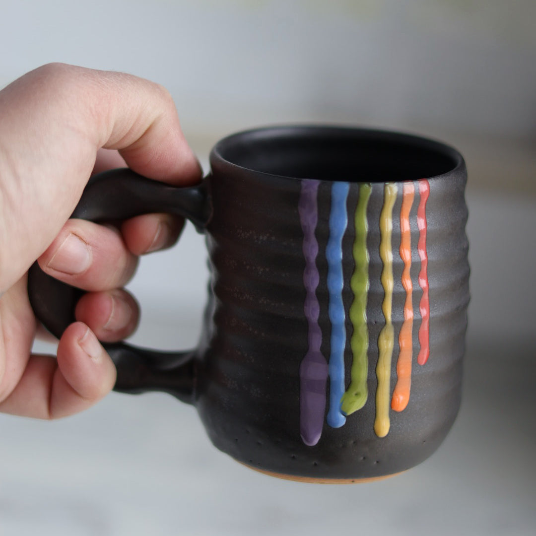 "Hand holding black ceramic companion mug with vibrant rainbow colored glaze drips cascading down the side, showing purple, blue, green, yellow, orange and red stripes"