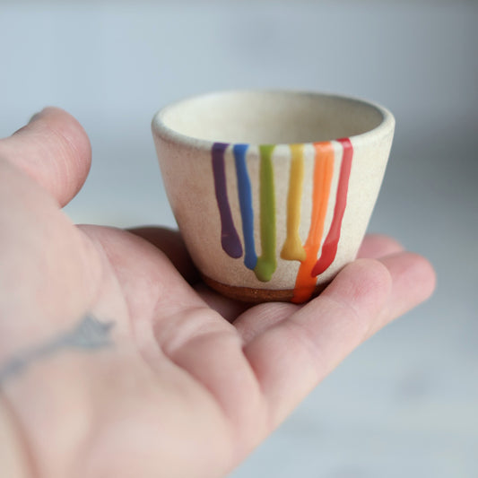 "Small cream ceramic bowl with rainbow drip glaze held in palm of hand, showing colorful striped pattern dripping from rim"