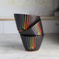 "Three black ceramic bowls stacked at angles showing rainbow drip glaze patterns with vibrant colored stripes against dark background"