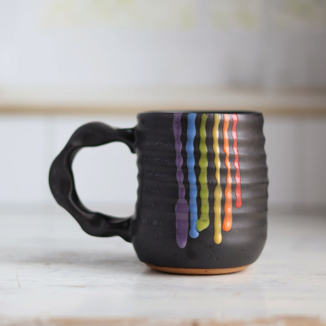 "Black ceramic companion mug with vibrant rainbow colored glaze drips cascading down the side, showing purple, blue, green, yellow, orange and red stripes"