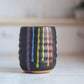 "Black ceramic wine cup with textured surface and rainbow drip glaze showing vibrant colored stripes"