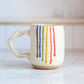 "Cream colored ceramic companion mug with handle featuring rainbow drip glaze pattern with purple, blue, green, yellow, orange and red stripes flowing down the side"