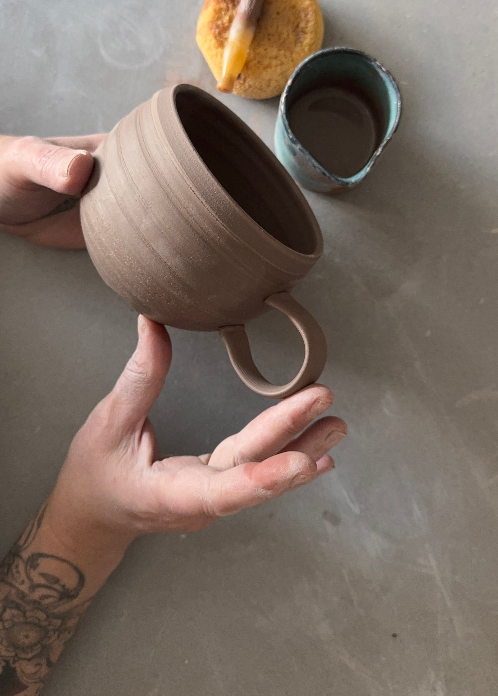 A person's hands hold a handmade, unglazed brown clay mug over a gray workbench, with pottery tools and a small blue cup in the background.