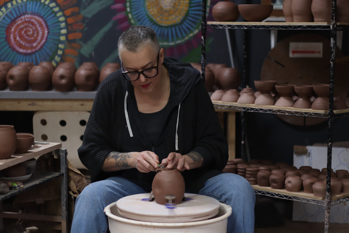 A person wearing glasses and a black hoodie sits at a pottery wheel, shaping a terracotta pot in a workshop filled with shelves of ceramic vessels.