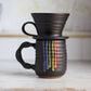 "Black ceramic companion mug with vibrant rainbow colored glaze drips cascading down the side, showing purple, blue, green, yellow, orange and red stripes, photographed with the black V60 coffee dripper"