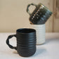 Black textured mug on a light surface with a blurred cream and green mug in the background.