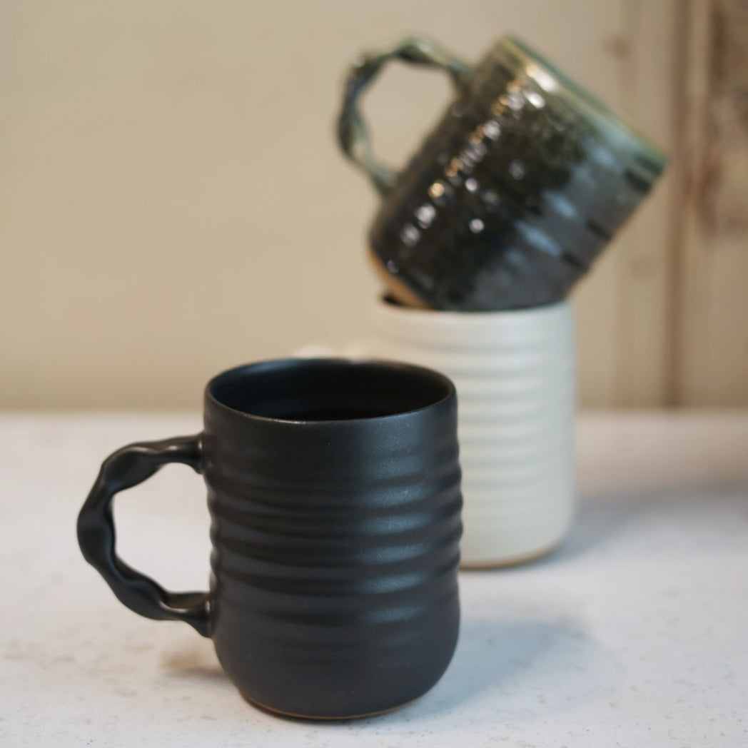 Black textured mug on a light surface with a blurred cream and green mug in the background.
