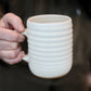 Person holding a cream ceramic mug with ribbed texture against a blurred background