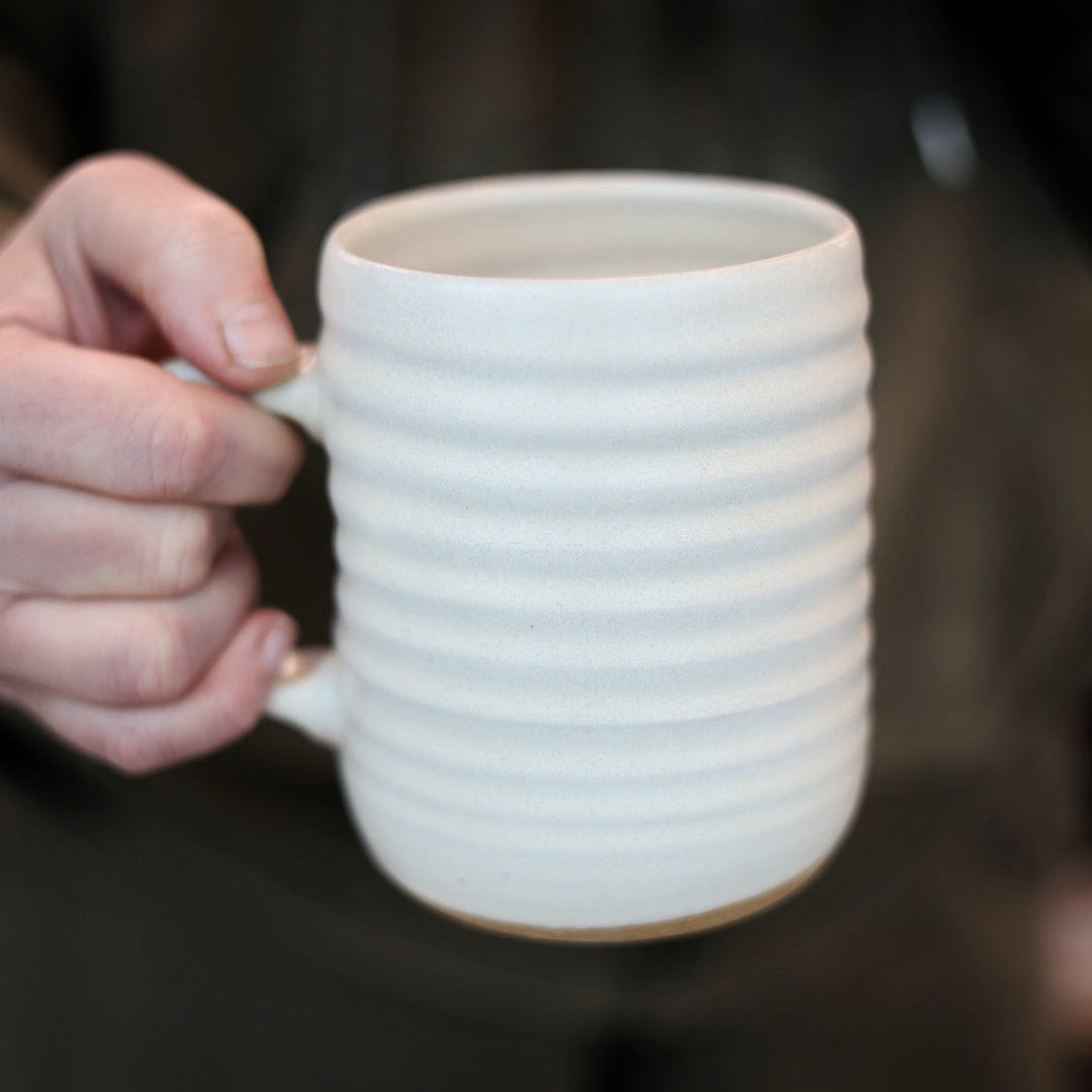 Person holding a cream ceramic mug with ribbed texture against a blurred background