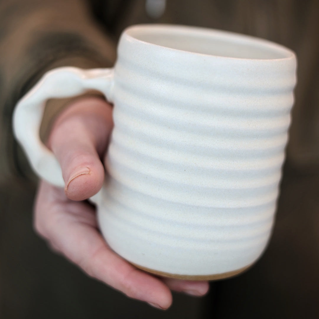 Hand holding a cream ceramic mug with textured design against a blurred background