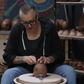 A person with short gray hair and glasses sits at a pottery wheel, shaping a small terracotta pot. Shelves in the background are filled with finished clay vessels and pottery pieces.