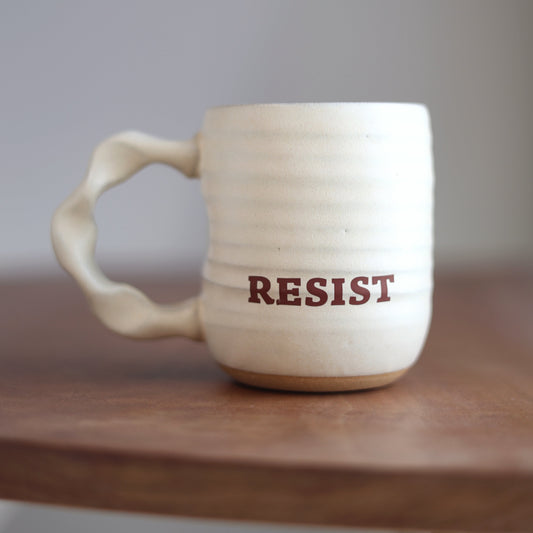 White ceramic mug with 'RESIST' printed on it, placed on a wooden surface.