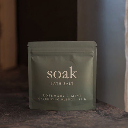 Green package of 'soak' bath salt with 'Rosemary + Mint' flavor on a dark surface.
