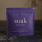 Purple package of 'soak' bath salt on a dark surface with a blurred background