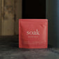Coral packet of 'soak' bath salt on a dark surface with a blurred background