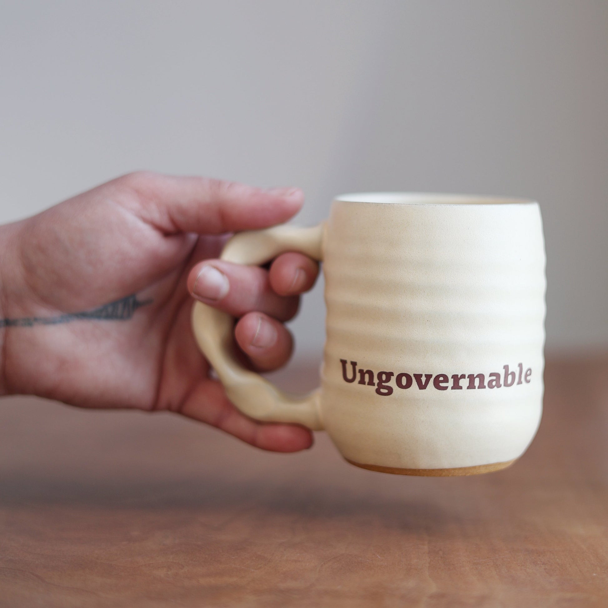 Hand holding a mug with 'Ungovernable' text on a wooden surface