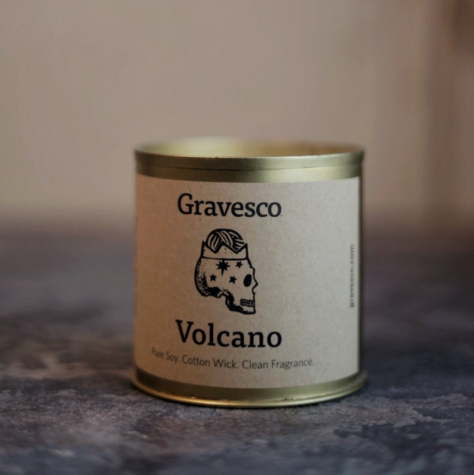 Candle in a tin labeled 'Gravesco Volcano' on a textured surface with a blurred background