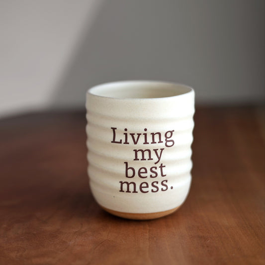 White ceramic mug with text 'Living my best mess' on a wooden surface.