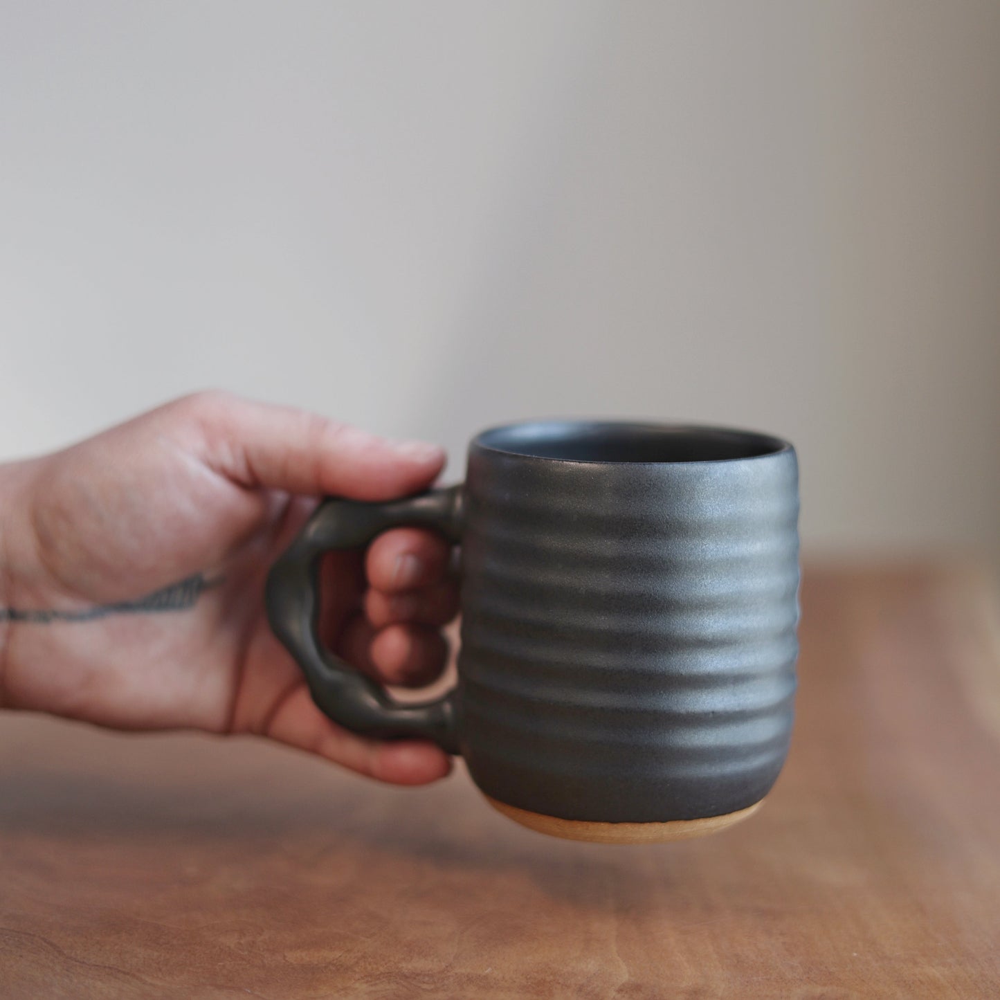 Hand holding a black ceramic mug on a wooden surface with a neutral background