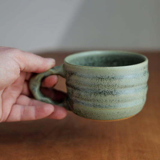 Hand holding a green ceramic mug on a wooden surface with a blurred background